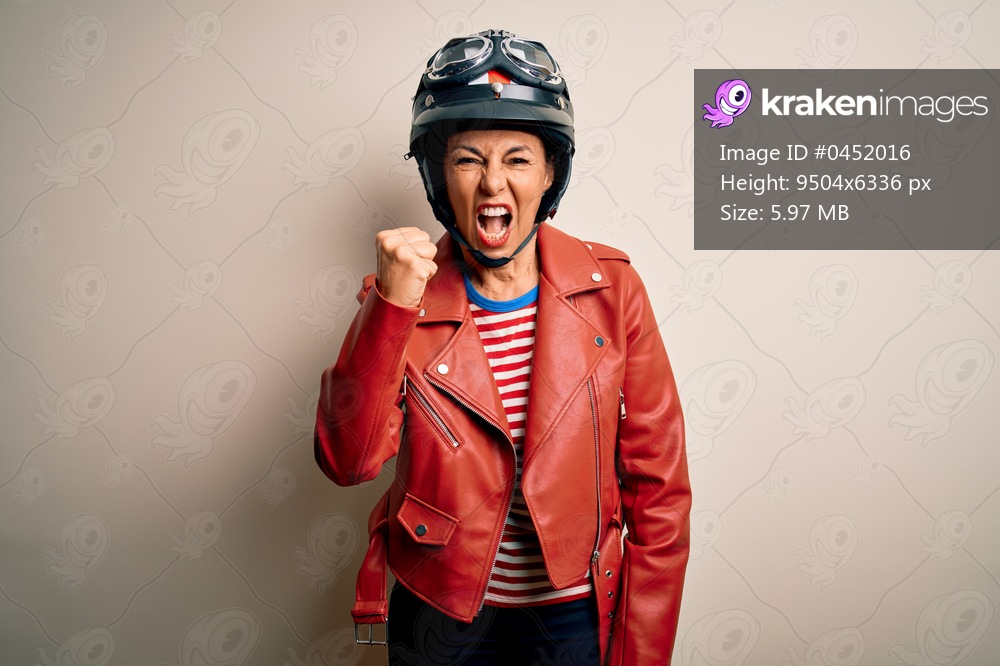 Middle age motorcyclist woman wearing motorcycle helmet and jacket over white background angry and mad raising fist frustrated and furious while shouting with anger. Rage and aggressive concept.