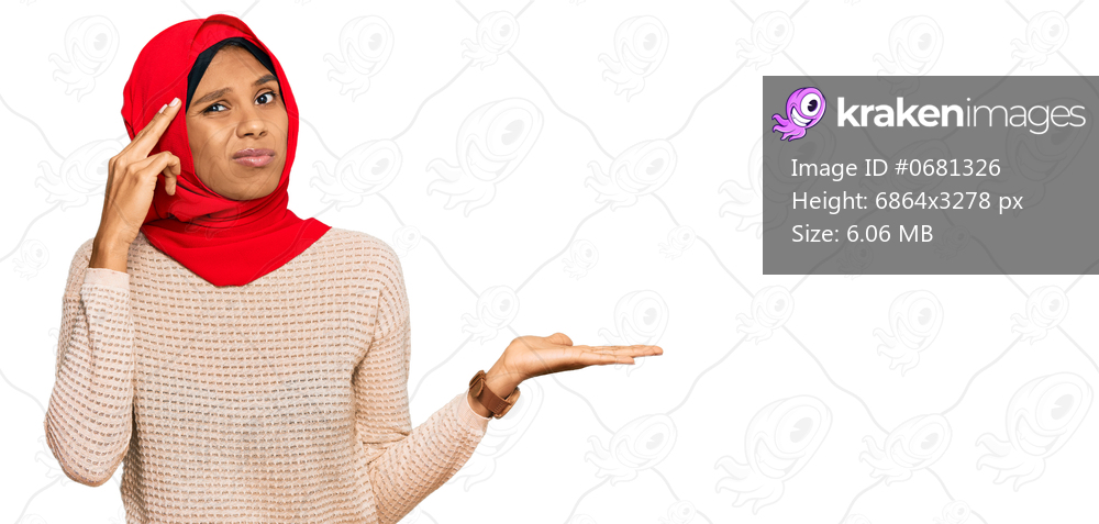 Young african american woman wearing traditional islamic hijab scarf confused and annoyed with open palm showing copy space and pointing finger to forehead. think about it. 