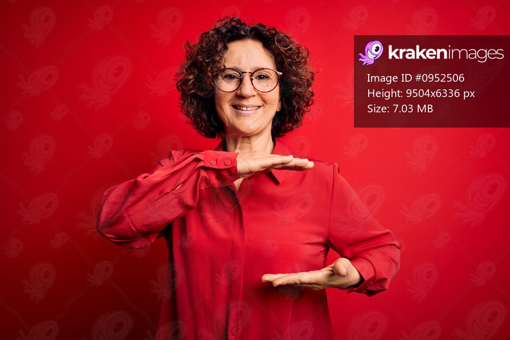 Middle age beautiful curly hair woman wearing casual shirt and glasses over red background gesturing with hands showing big and large size sign, measure symbol. Smiling looking at the camera. Measuring concept.