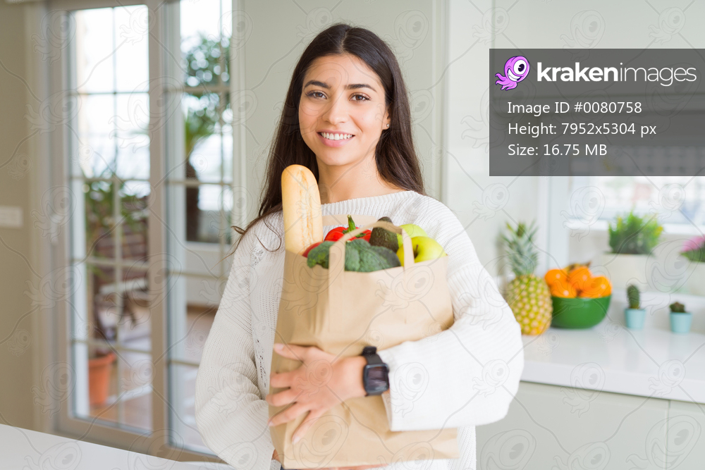 Beautiful young woman smiling holding a paper bag full of groceries at the kitchen