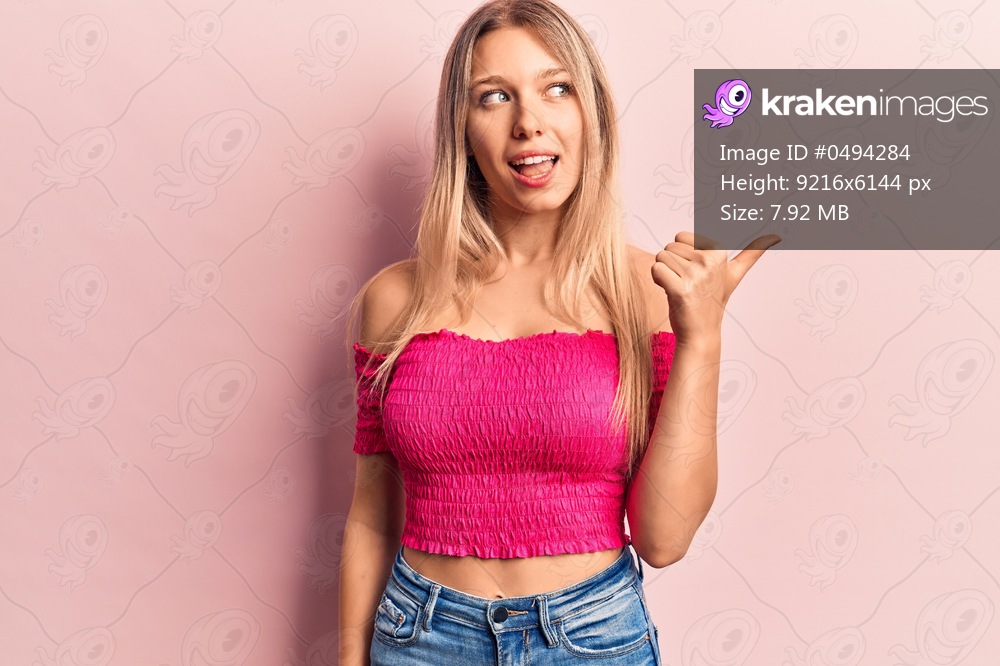 Young blonde woman wearing casual clothes pointing thumb up to the side smiling happy with open mouth 