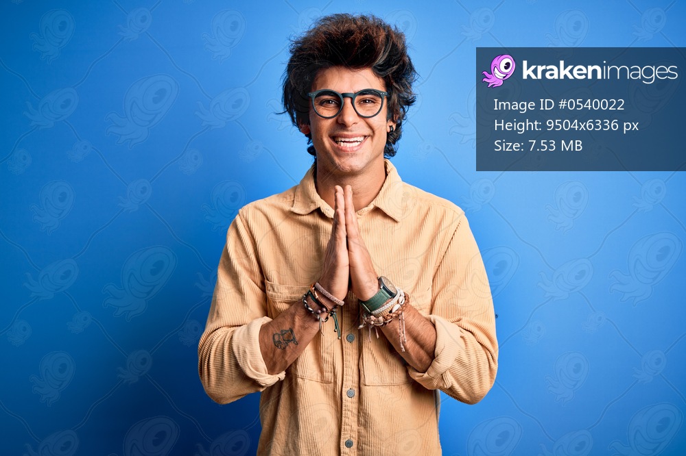 Young handsome man wearing casual shirt standing over isolated blue background praying with hands together asking for forgiveness smiling confident.
