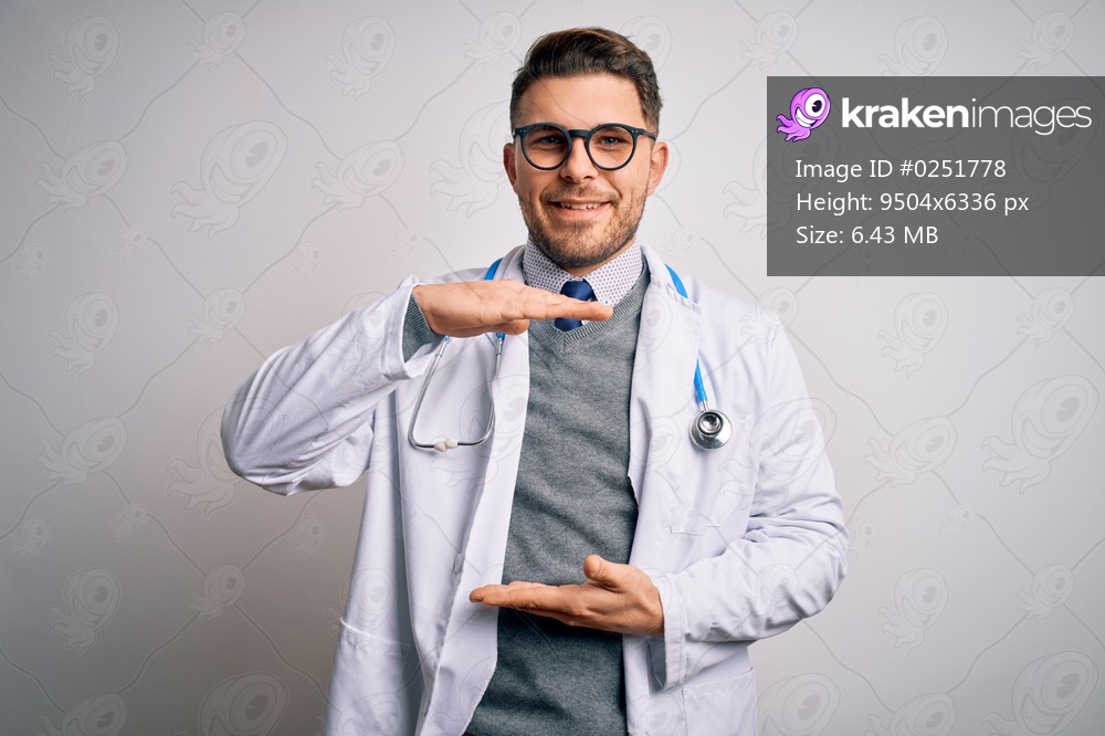 Young doctor man with blue eyes wearing medical coat and stethoscope over isolated background gesturing with hands showing big and large size sign, measure symbol. Smiling looking at the camera. Measuring concept.