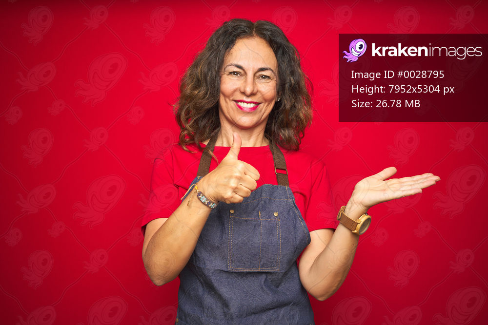 Middle age senior woman wearing apron uniform over red isolated background Showing palm hand and doing ok gesture with thumbs up, smiling happy and cheerful