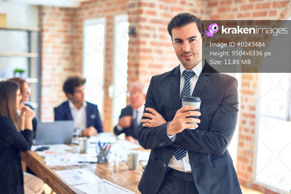 Group of business workers smiling happy and confident working together in a meeting. One of them, standing with smile on face looking at camera drinking coffee at the office.