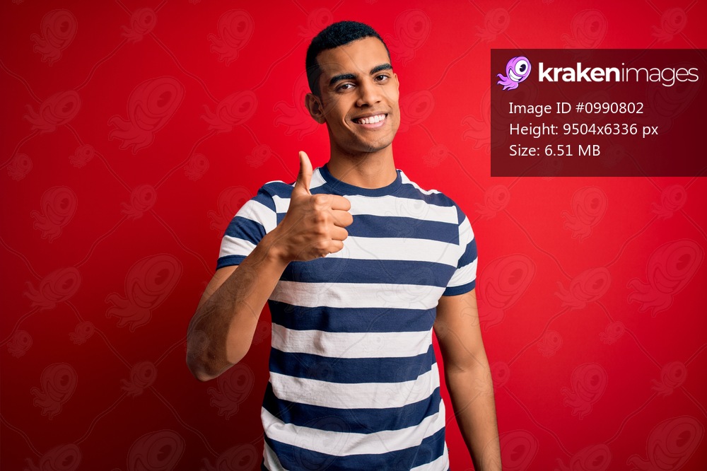Handsome african american man wearing casual striped t-shirt standing over red background doing happy thumbs up gesture with hand. Approving expression looking at the camera showing success.