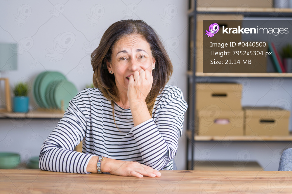 Middle age senior woman sitting at the table at home looking stressed and nervous with hands on mouth biting nails. Anxiety problem.