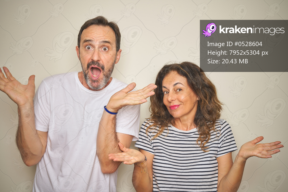 Beautiful middle age couple together standing over isolated white background clueless and confused expression with arms and hands raised. Doubt concept.
