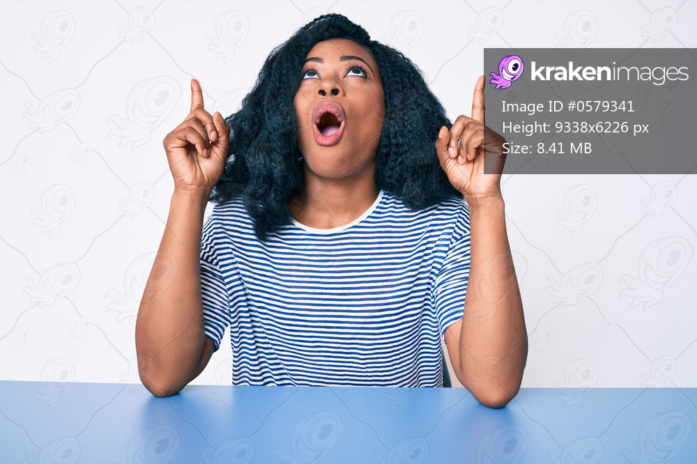 Beautiful african woman wearing casual clothes sitting on the table amazed and surprised looking up and pointing with fingers and raised arms. 