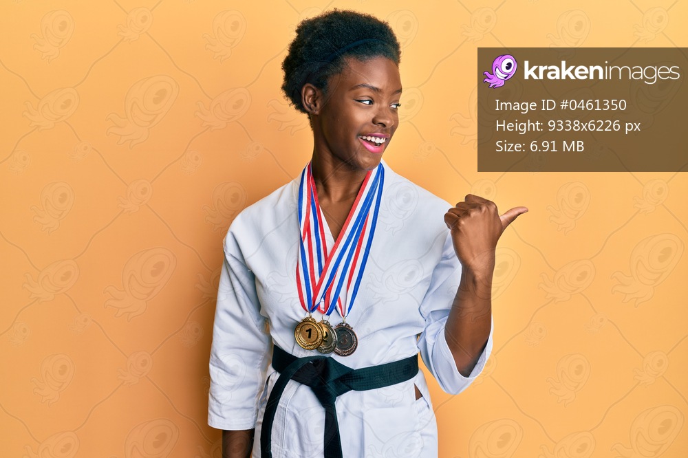 Young african american girl wearing karate kimono and black belt smiling with happy face looking and pointing to the side with thumb up. 