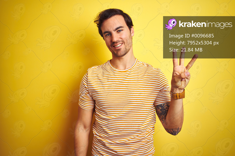 Young handsome man with tattoo wearing striped t-shirt over isolated yellow background showing and pointing up with fingers number three while smiling confident and happy.