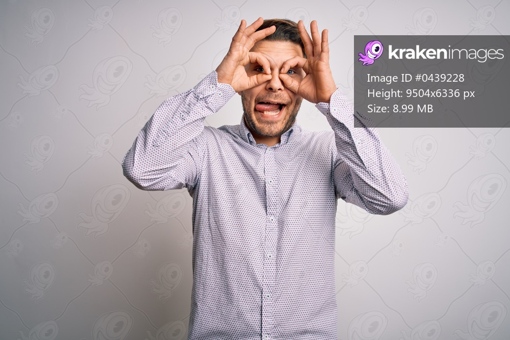 Young business man with blue eyes standing over isolated background doing ok gesture like binoculars sticking tongue out, eyes looking through fingers. Crazy expression.