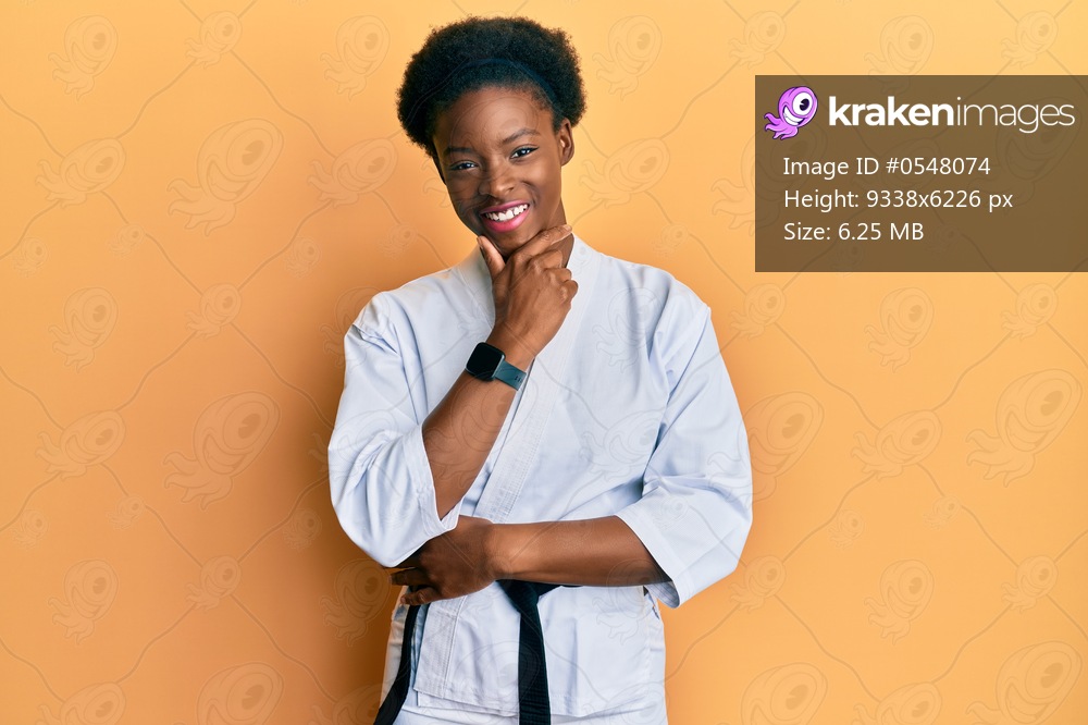 Young african american girl wearing karate kimono and black belt looking confident at the camera smiling with crossed arms and hand raised on chin. thinking positive. 