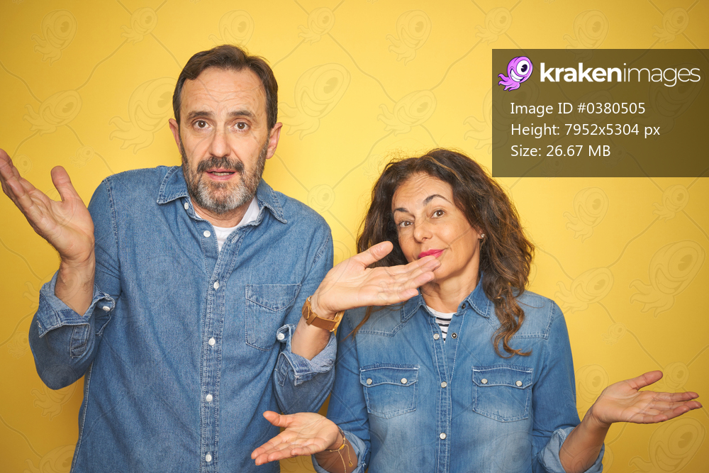 Beautiful middle age couple together standing over isolated yellow background clueless and confused expression with arms and hands raised. Doubt concept.