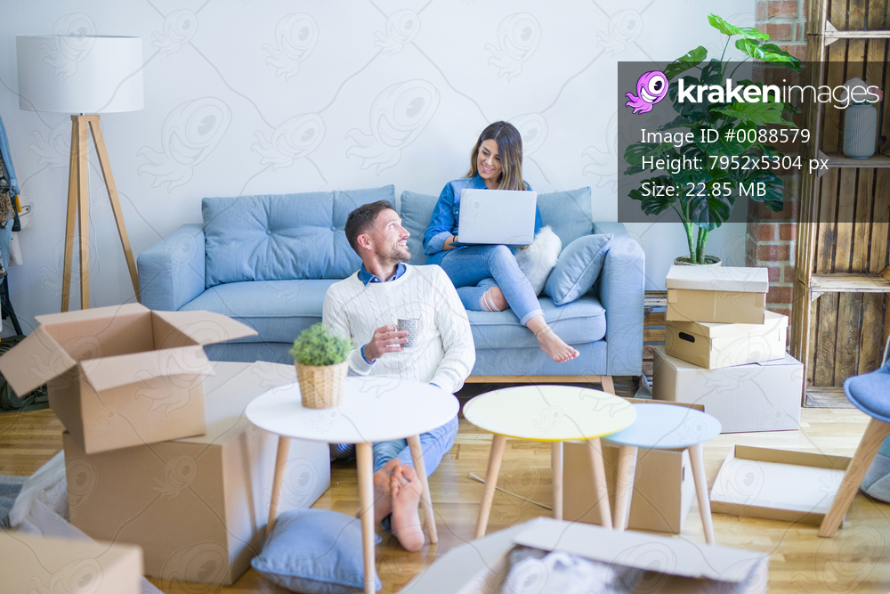 Young beautiful couple sitting on the sofa drinking cup of coffee using laptop at new home around cardboard boxes