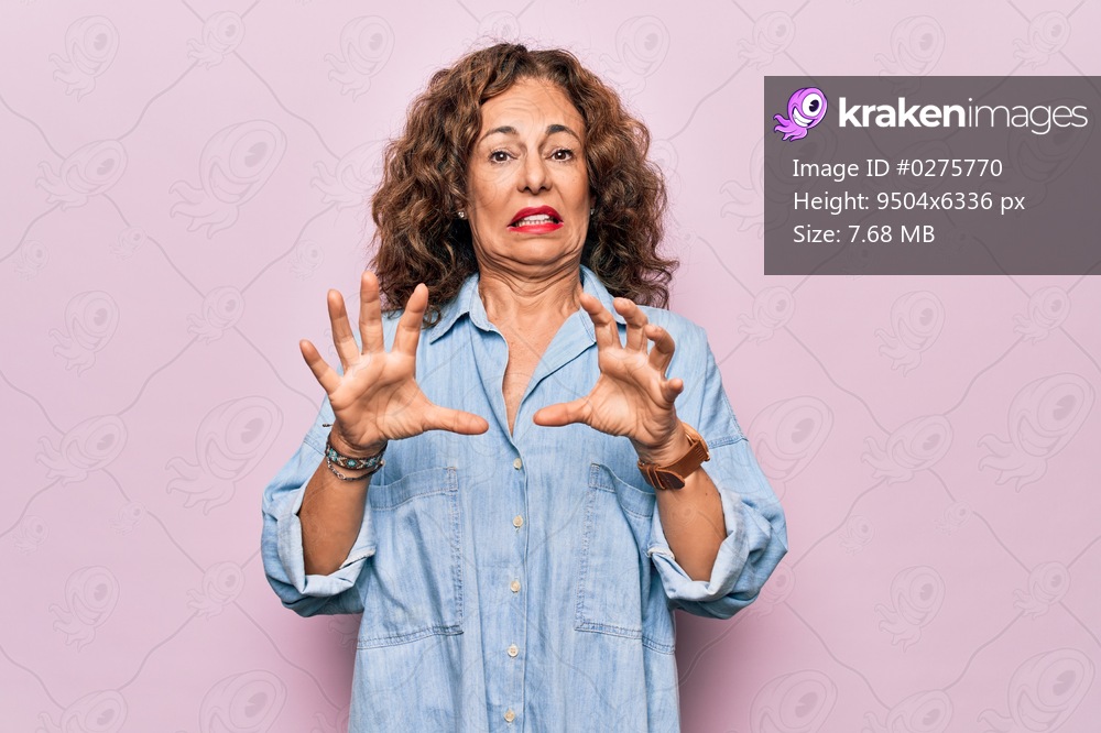 Middle age beautiful woman wearing casual denim shirt standing over pink background afraid and terrified with fear expression stop gesture with hands, shouting in shock. Panic concept.