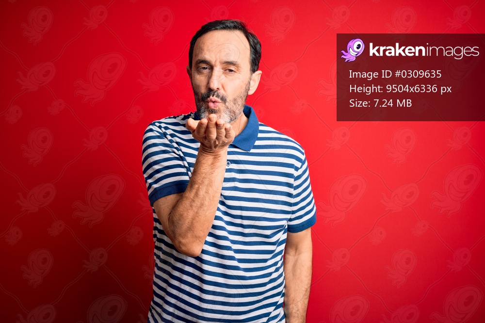 Middle age hoary man wearing casual striped polo standing over isolated red background looking at the camera blowing a kiss with hand on air being lovely and sexy. Love expression.