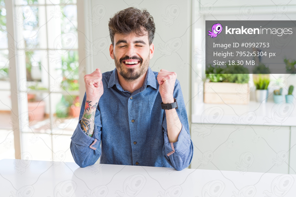 Young man wearing casual shirt sitting on white table excited for success with arms raised celebrating victory smiling. Winner concept.