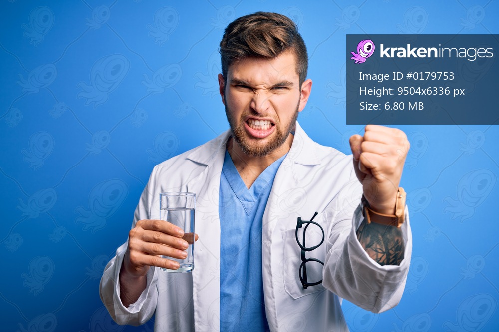 Young blond doctor man with beard and blue eyes wearing coat drinking glass of water annoyed and frustrated shouting with anger, crazy and yelling with raised hand, anger concept
