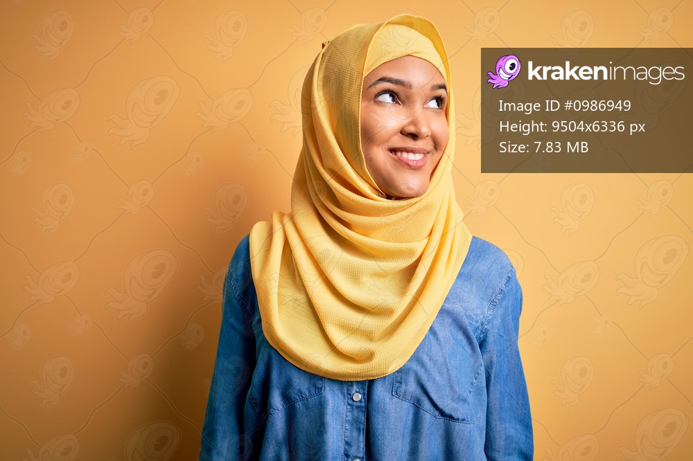 Young beautiful woman with curly hair wearing arab traditional hijab over yellow background smiling looking to the side and staring away thinking.