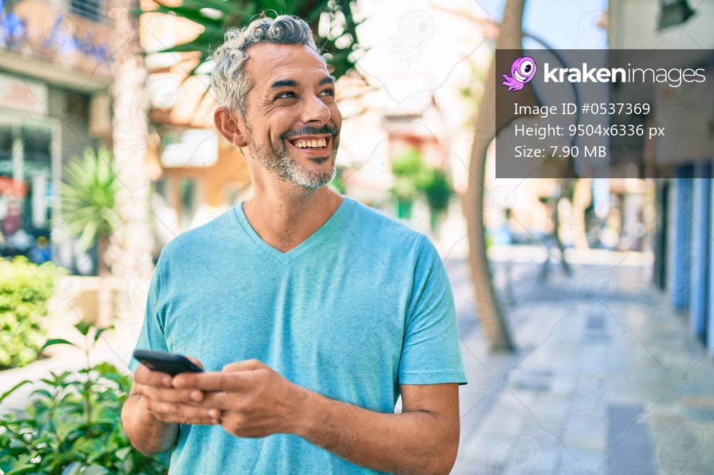 Middle age grey-haired man smiling happy using smartphone walking at street of city.