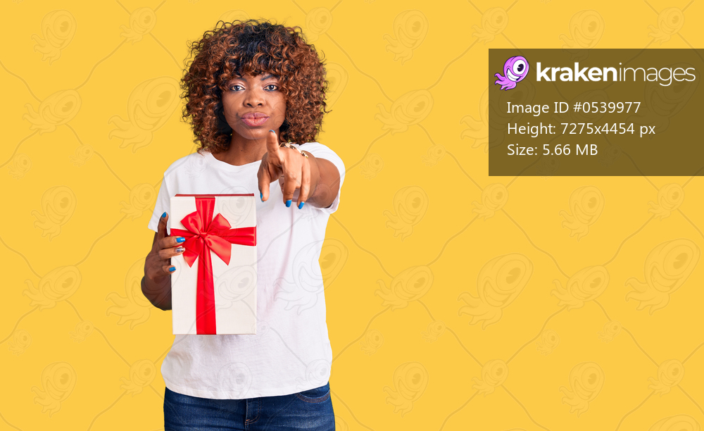 Young african american woman holding gift pointing with finger to the camera and to you, confident gesture looking serious 