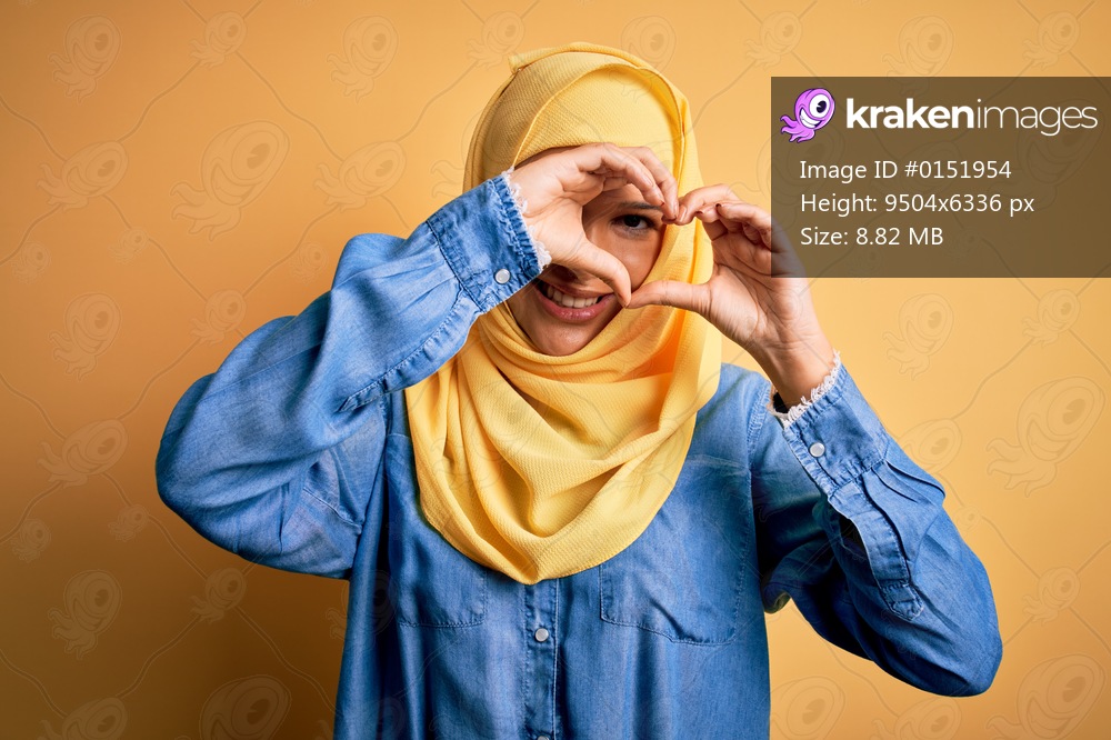 Young beautiful woman with curly hair wearing arab traditional hijab over yellow background Doing heart shape with hand and fingers smiling looking through sign