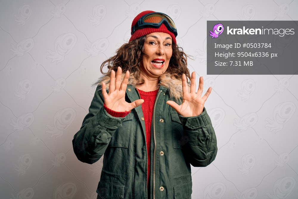 Middle age skier woman wearing snow sportswear and ski goggles over white background afraid and terrified with fear expression stop gesture with hands, shouting in shock. Panic concept.