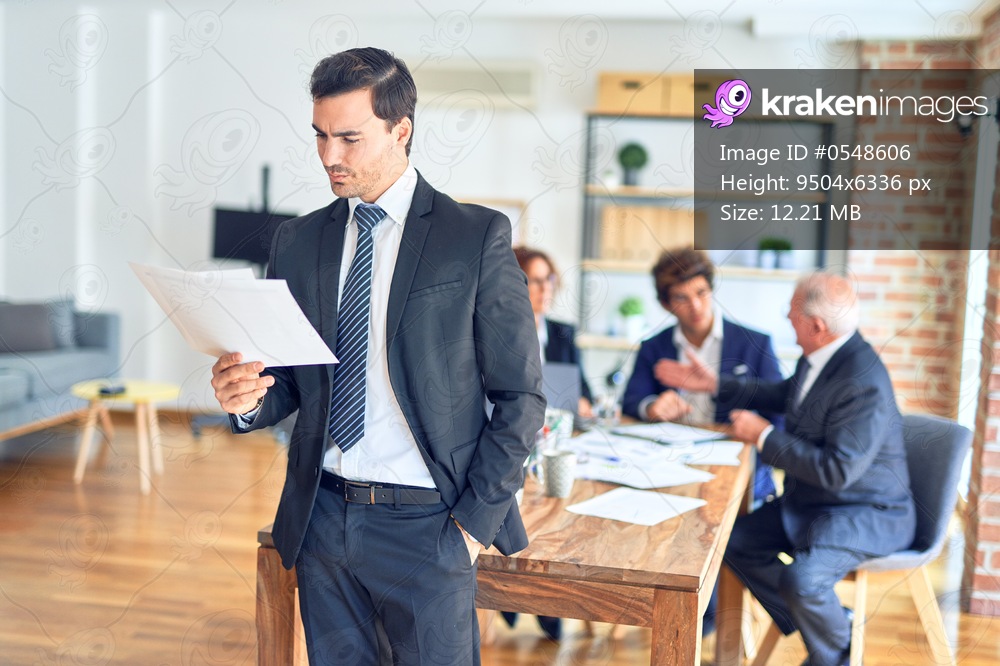 Group of business workers smiling happy and confident working together in a meeting. One of them, standing with smile on face reading documents at the office.