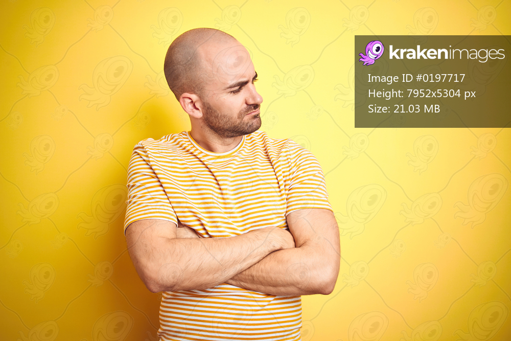 Young bald man with beard wearing casual striped t-shirt over yellow isolated background looking to the side with arms crossed convinced and confident