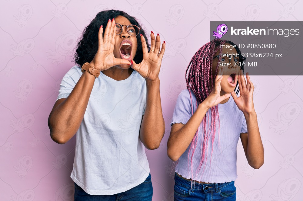 Beautiful african american mother and daughter wearing casual clothes and glasses shouting angry out loud with hands over mouth 