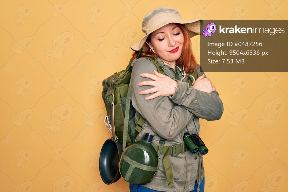 Young redhead backpacker woman hiking wearing backpack and hat over yellow background Hugging oneself happy and positive, smiling confident. Self love and self care