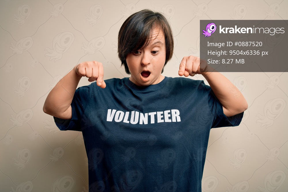 Young down syndrome volunteer woman wearing social care charity t-shirt Pointing down with fingers showing advertisement, surprised face and open mouth