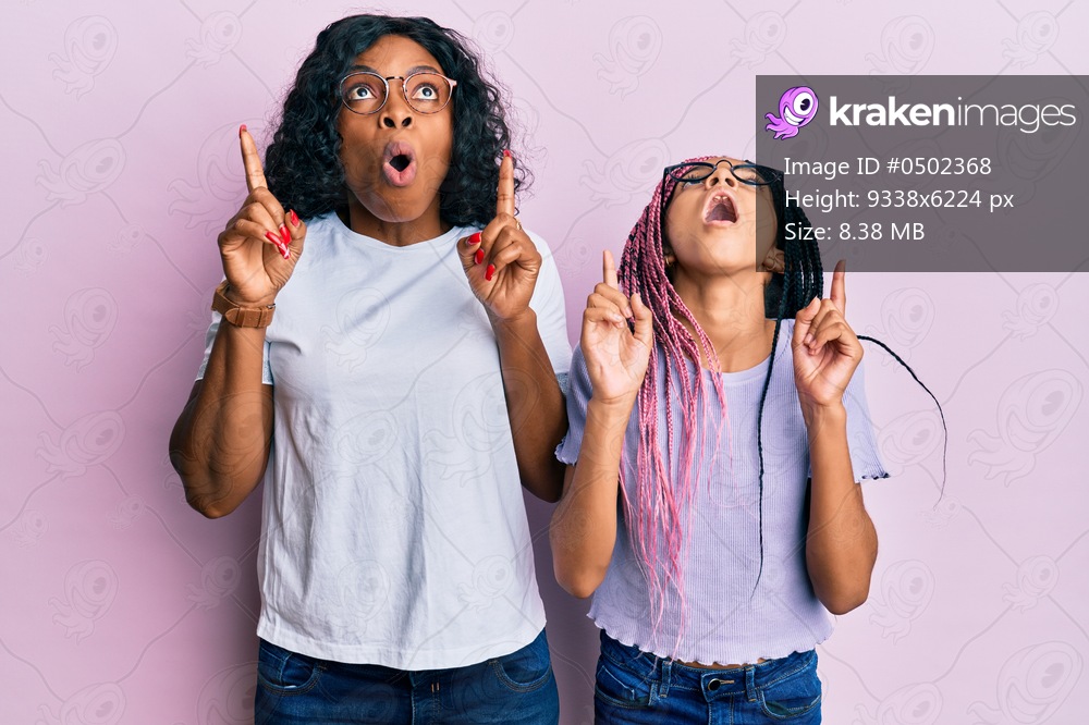 Beautiful african american mother and daughter wearing casual clothes and glasses amazed and surprised looking up and pointing with fingers and raised arms. 