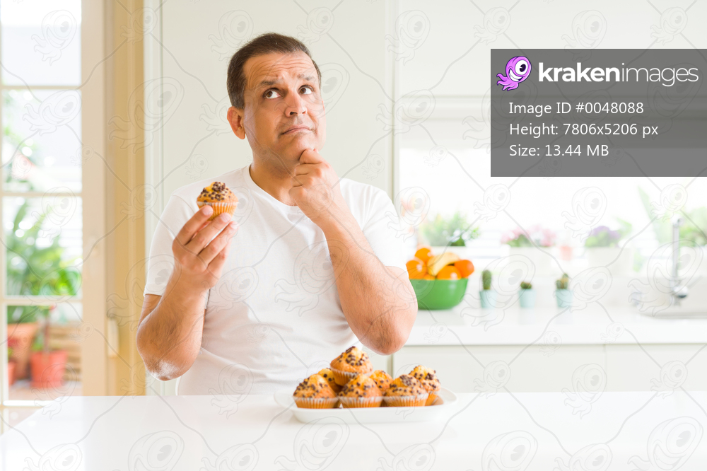 Middle age man eating chocolate chip muffins at home serious face thinking about question, very confused idea