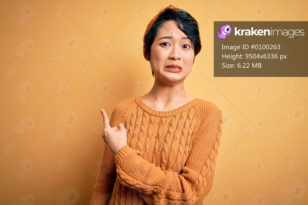 Young beautiful asian girl wearing casual sweater and diadem standing over yellow background Pointing aside worried and nervous with forefinger, concerned and surprised expression