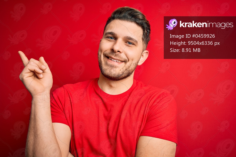 Young man with blue eyes wearing casual t-shirt over red isolated background with a big smile on face, pointing with hand and finger to the side looking at the camera.