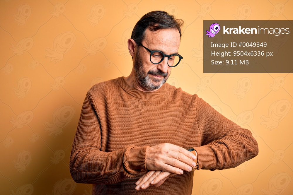 Middle age hoary man wearing brown sweater and glasses over isolated yellow background Checking the time on wrist watch, relaxed and confident