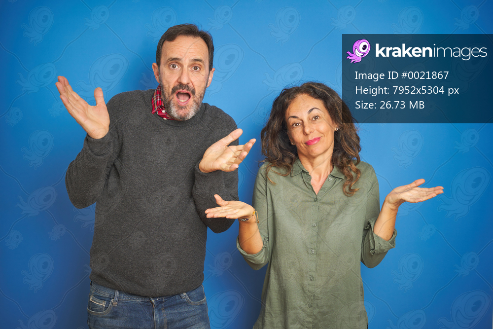 Beautiful middle age couple together standing over isolated blue background clueless and confused expression with arms and hands raised. Doubt concept.