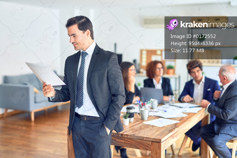 Group of business workers smiling happy and confident working together in a meeting. One of them, standing with smile on face reading documents at the office.