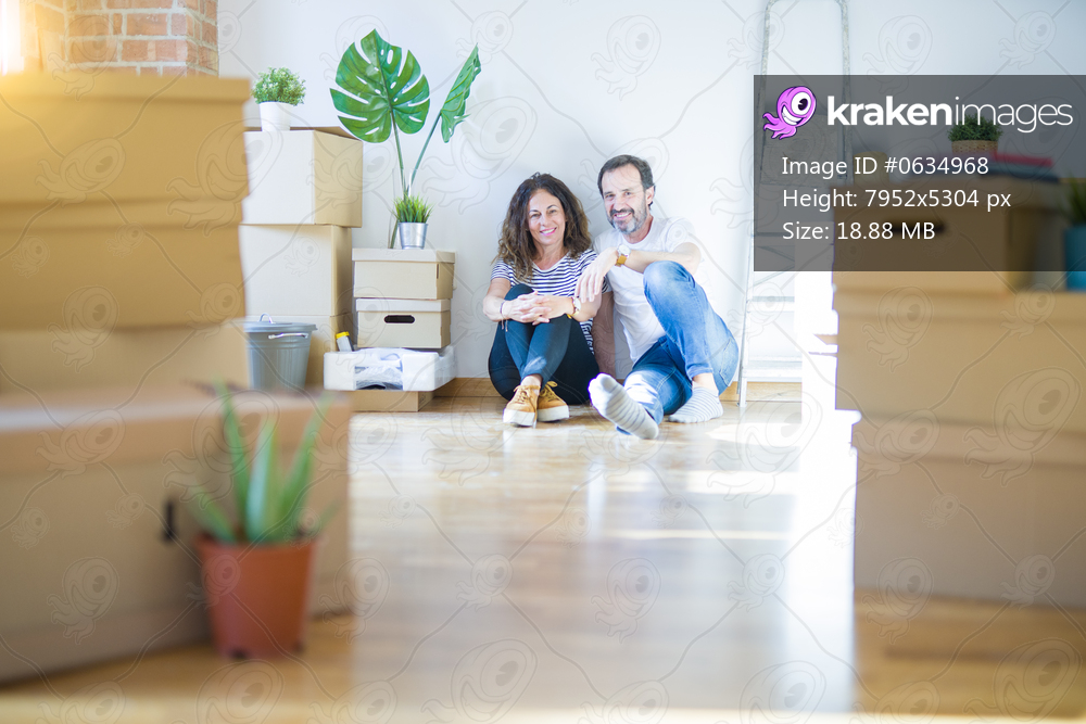 Middle age senior romantic couple in love sitting on the apartment floor with cardboard boxes around and smiling happy for moving to a new home