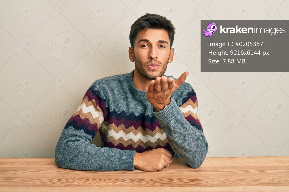 Young handsome man wearing casual sweater sitting on the table looking at the camera blowing a kiss with hand on air being lovely and sexy. love expression. 
