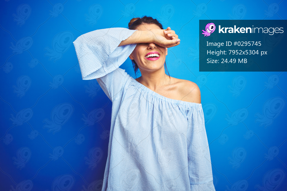 Young beautiful woman wearing bun hairstyle over blue isolated background covering eyes with arm smiling cheerful and funny. Blind concept.