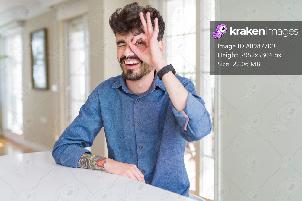 Young man wearing casual shirt sitting on white table doing ok gesture with hand smiling, eye looking through fingers with happy face.
