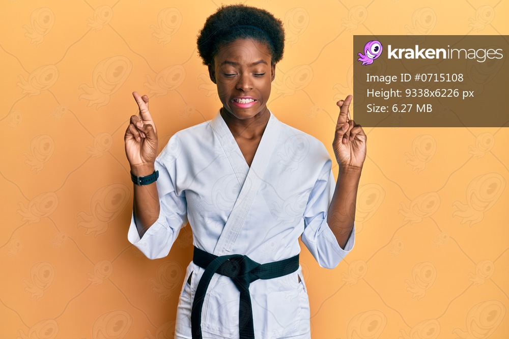 Young african american girl wearing karate kimono and black belt gesturing finger crossed smiling with hope and eyes closed. luck and superstitious concept. 