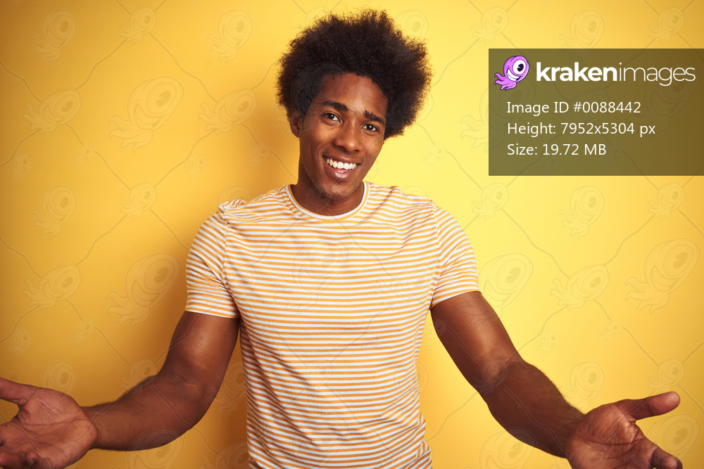 American man with afro hair wearing striped t-shirt standing over isolated yellow background smiling cheerful with open arms as friendly welcome, positive and confident greetings