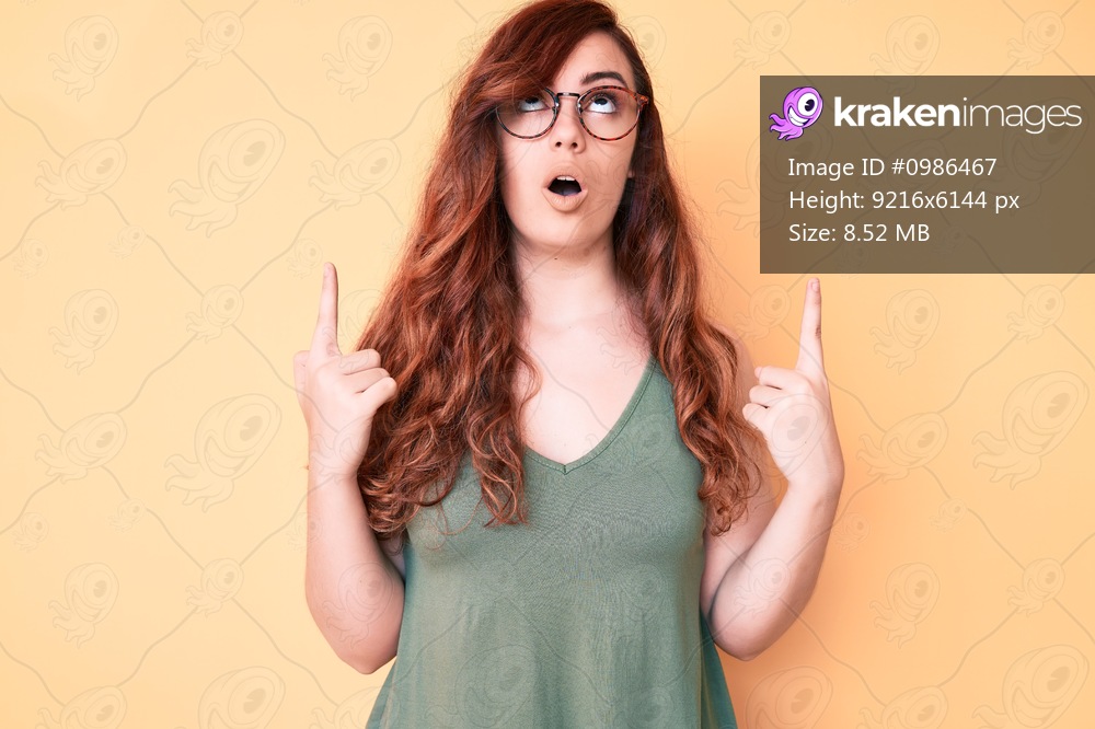 Young beautiful woman wearing casual clothes and glasses amazed and surprised looking up and pointing with fingers and raised arms. 
