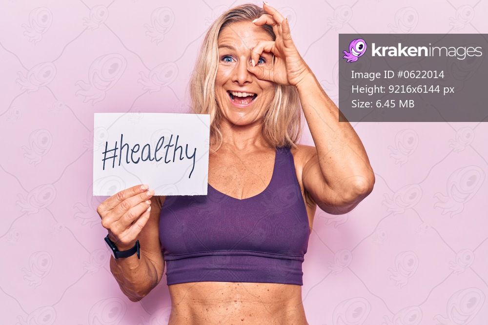 Middle age caucasian blonde woman wearing sportswear holding healthy banner smiling happy doing ok sign with hand on eye looking through fingers 