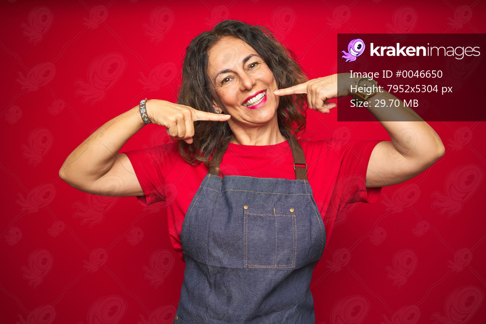 Middle age senior woman wearing apron uniform over red isolated background smiling cheerful showing and pointing with fingers teeth and mouth. Dental health concept.