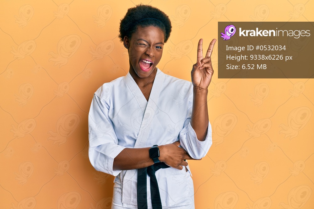 Young african american girl wearing karate kimono and black belt smiling with happy face winking at the camera doing victory sign. number two. 